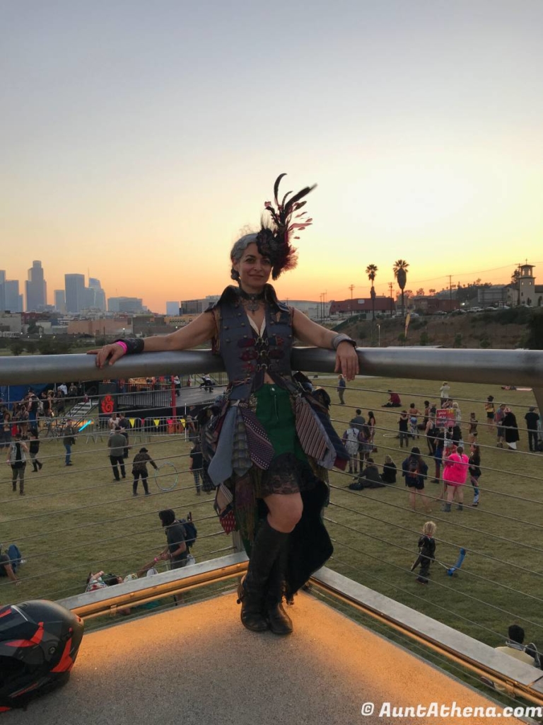 Aunt Athena with feathers in her hair wearing a tie dress standing at a railing with a park full of people behind her and the downtown skyline behind that
